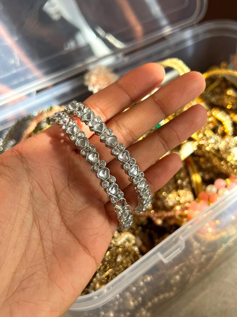May include: Two silver bangles with heart-shaped clear stones are held in a hand. The bangles are in focus, with other jewelry visible in the background. The jewelry is stored in a clear plastic container.