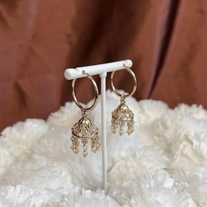 May include: A pair of gold-coloured hoop earrings with a bell-shaped charm. The charm is adorned with small, light-coloured beads and decorative detailing. The earrings are displayed on a white stand against a brown background.