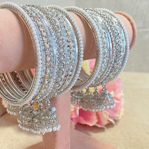 May include: A set of silver-toned bangles featuring intricate metallic patterns, rows of small white faux pearls, and clear crystals. Each bangle is adorned with multiple dangling silver-toned bell-shaped charms embellished with tiny white faux pearls.