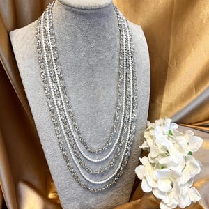 Puede incluir: Un collar plateado de múltiples cadenas con una cadena de perlas en el centro. El collar se muestra en un busto de maniquí blanco.