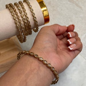 May include: A set of gold bangles with clear rhinestones. The bangles are stacked on a wooden display stand. One bangle is worn on a person's wrist.