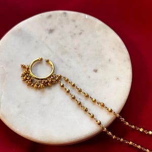 May include: A gold-coloured nose ring with a beaded chain and dangling accents. The nose ring is circular with a small cluster of beads on the bottom and two beaded chains. The jewellery is displayed on a white marble surface against a red background.