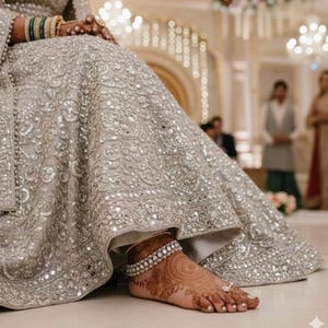 May include: A silver wedding dress with intricate embroidery and mirror embellishments. The bride's foot is adorned with a silver anklet and henna designs. The dress is floor-length, with a detailed pattern.