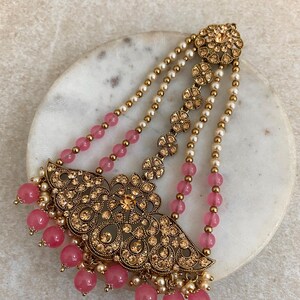 Puede incluir: Un tocado dorado ornamentado con intrincados filigranas y acentos brillantes. Cuentas rosas y perlas de imitación cuelgan del diseño. El tocado es probablemente para una ocasión especial, como una boda o un evento formal.