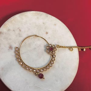 May include: A gold-coloured nose ring with a circular design. It features small, clear stones and a cluster of red gemstones. A small red bead hangs from the bottom, and a delicate chain extends from the top.