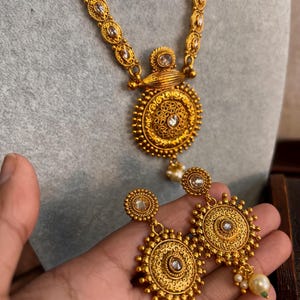 May include: A gold-coloured necklace and earring set. The necklace has a large pendant with a central stone and detailed patterns. The earrings reflect the pendant's design, with dangling pearls. The set is shown against a neutral backdrop.