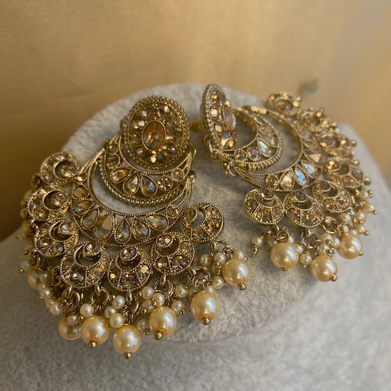 Peut inclure: Une paire de boucles d'oreilles en or avec des motifs complexes et des perles pendantes. Les boucles d'oreilles pr&eacute;sentent une forme de croissant de lune avec plusieurs couches d'embellissements.