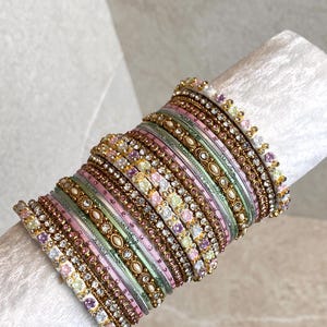 Puede incluir: Una pila de múltiples brazaletes que presentan una variedad de diseños, incluyendo bandas de oro metálico, bandas texturizadas en rosa pastel, verde y blanco, y elementos decorativos como pequeños cristales incrustados y diminutos patrones florales.