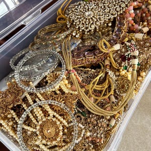 May include: A clear plastic storage container overflowing with an assortment of ornate jewelry. The jewelry includes necklaces, bracelets, and earrings in gold, silver, and pearl tones. Many pieces are embellished with rhinestones and colorful accents.