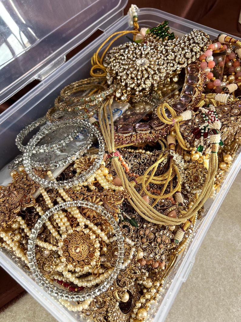 May include: A clear plastic storage container overflowing with an assortment of ornate jewelry. The jewelry includes necklaces, bracelets, and earrings in gold, silver, and pearl tones. Many pieces are embellished with rhinestones and colorful accents.