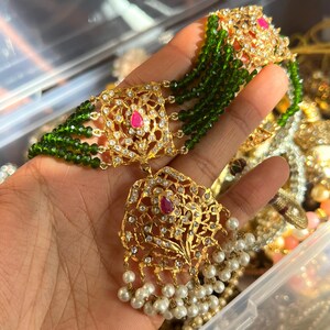 May include: Gold-toned necklace with intricate filigree design, adorned with clear crystals, a central pink gemstone, and dangling white pearls. The necklace features strands of green beads, creating a layered effect. A luxurious piece of jewelry.
