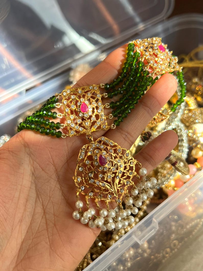 May include: Gold-toned necklace with intricate filigree design, adorned with clear crystals, a central pink gemstone, and dangling white pearls. The necklace features strands of green beads, creating a layered effect. A luxurious piece of jewelry.