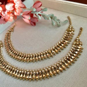 May include: Two gold-colored anklets with a teardrop design, featuring small, clear accents. The anklets are arranged on a light-colored surface, with a sprig of pink flowers in the background. The jewelry is likely for special occasions.