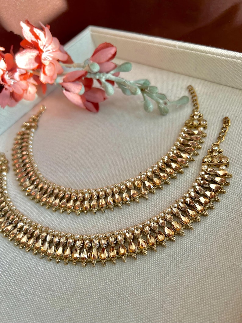 May include: Two gold-colored anklets with a teardrop design, featuring small, clear accents. The anklets are arranged on a light-colored surface, with a sprig of pink flowers in the background. The jewelry is likely for special occasions.