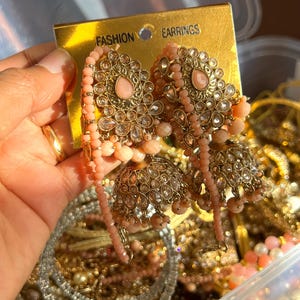 May include: Ornate gold-toned earrings with pink gemstones and dangling beads. The earrings have a floral design and are labeled "FASHION EARRINGS" on a gold tag. The background shows other jewelry pieces.