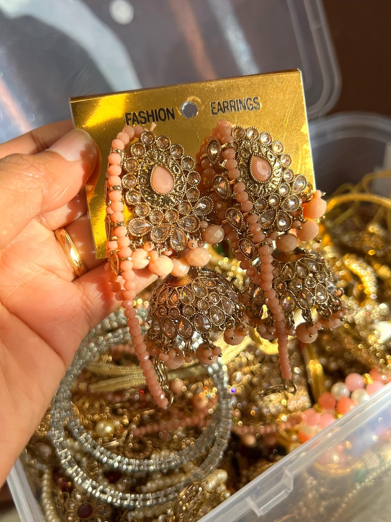 May include: Ornate gold-toned earrings with pink gemstones and dangling beads. The earrings have a floral design and are labeled "FASHION EARRINGS" on a gold tag. The background shows other jewelry pieces.