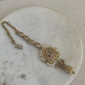 May include: Gold-toned nose ring with a geometric design, featuring a clear stone surrounded by smaller stones. The ring is attached to a chain with a hook closure and has a pearl and tassel drop.
