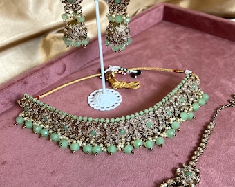 May include: A gold and green Indian bridal jewellery set featuring a choker necklace, earrings, and a maang tikka. The necklace is decorated with intricate floral patterns and green beads. The earrings are drop style with a similar design to the necklace.