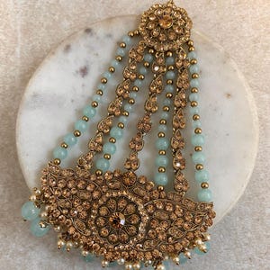 May include: An ornate gold-toned hair accessory with intricate floral and leaf patterns. It features light blue beads and small white pearls. The design includes a central flower-shaped element and dangling beaded strands.