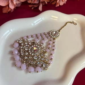 May include: An ornate, gold-toned headpiece with clear crystals and light pink beads. The headpiece features a central, floral-inspired design and a delicate chain for securing to the hair. The piece is displayed on a white surface.