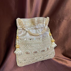 May include: Cream-coloured drawstring bag with detailed beadwork and mirror embellishments. The bag has a pearl beaded handle and decorative tassels. The design incorporates a detailed pattern of beads and small mirrors, creating a luxurious appearance.