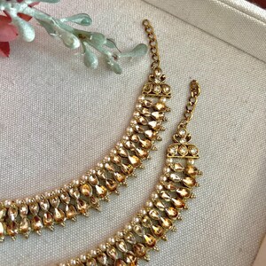 May include: Close-up of two gold-toned necklaces. Each necklace features a row of teardrop-shaped amber-colored stones and small white pearls. The necklaces have a delicate chain and ornate clasp. The jewelry is displayed on a neutral-toned surface.