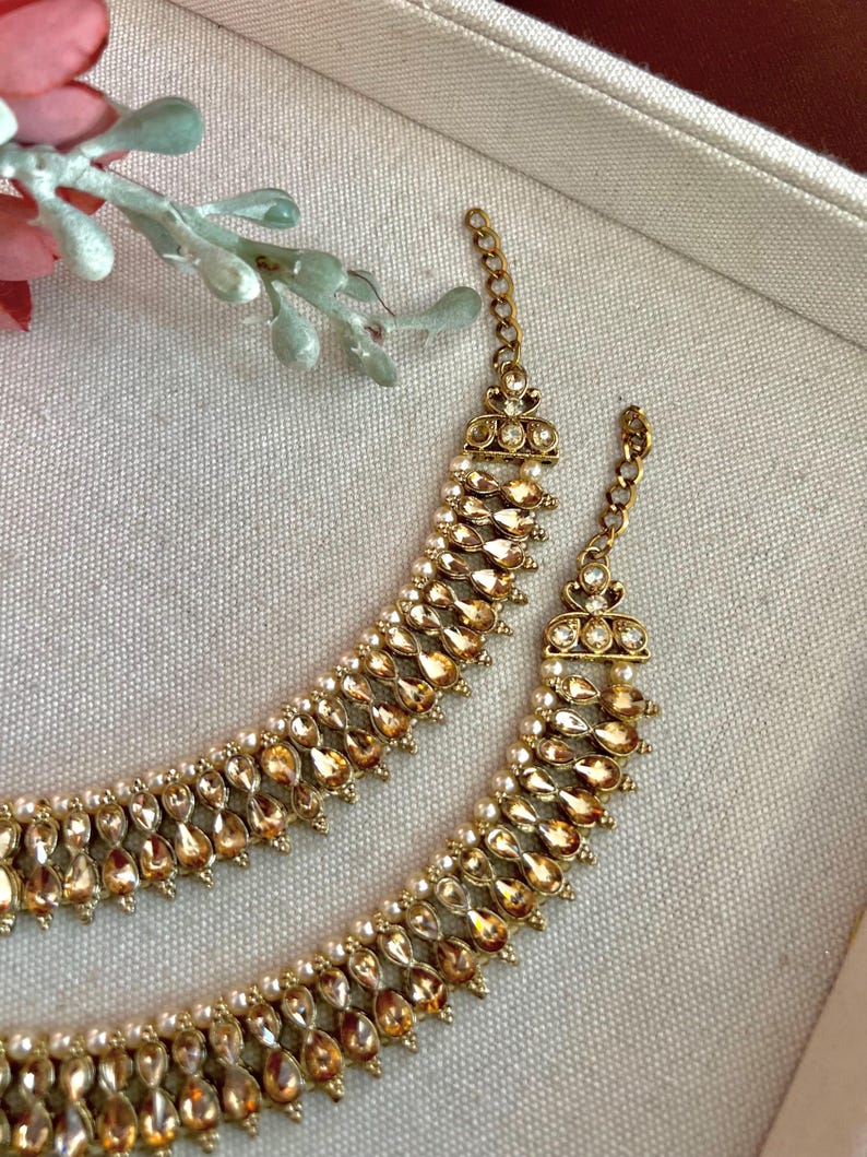 May include: Close-up of two gold-toned necklaces. Each necklace features a row of teardrop-shaped amber-colored stones and small white pearls. The necklaces have a delicate chain and ornate clasp. The jewelry is displayed on a neutral-toned surface.