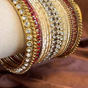 May include: A set of stacked bangles featuring a combination of gold-tone metal, white enamel bands, and rows of clear rhinestones. The design includes thin red accents and textured gold bead detailing along the edges of the circular bracelets.