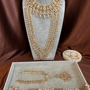 May include: An elaborate gold-toned necklace and matching jewelry set. The necklace features intricate floral and dangling designs. The set includes bracelets and additional necklaces, all adorned with sparkling accents and pearls. The jewelry is displayed on a gray mannequin and tray.