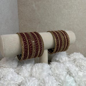 Peut inclure: Un ensemble de deux piles de bracelets rigides marron, chacun composé d'une combinaison de bandes marron unies et de bandes dorées ornées de petits ornements circulaires réfléchissants. Les bracelets sont disposés sur un présentoir cylindrique.
