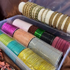 May include: A clear plastic storage box containing multiple sets of bangles. One row features gold-toned bangles with small white beads. The other rows contain stacks of solid-colored bangles in magenta, light pink, dark red, green, black, teal, and yellow.