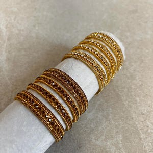 May include: A set of eight stacked bangles featuring a metallic gold finish. The top four bangles are adorned with clear white crystals, while the bottom four bangles are embellished with dark brown crystals, all set in a repeating pattern.
