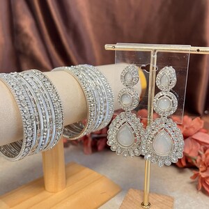 May include: A silver jewellery set comprising chandelier earrings and several bangles. The earrings have a teardrop shape with a large, round, white stone and are encrusted with small, clear crystals. The bangles are silver and decorated with clear crystals.