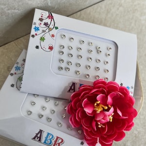 Puede incluir: Dos cajas blancas con ventanas de plástico transparente que contienen pequeños diamantes de imitación redondos plateados. Las cajas están etiquetadas con "ABR Bindi". Una flor rosa está en primer plano.