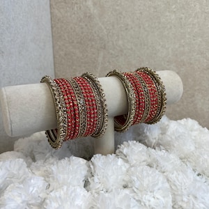 May include: A set of two matching bangle bracelets featuring alternating bands of bright red thread and metallic gold-toned wire. Each bracelet is adorned with small, clear, reflective stones embedded within the gold-toned metal sections.