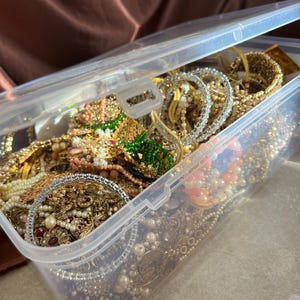 May include: A clear plastic storage container filled with various jewelry pieces. The jewelry includes gold-toned bracelets, necklaces, and earrings, adorned with pearls, beads, and gemstones. The container is partially open, revealing the contents.