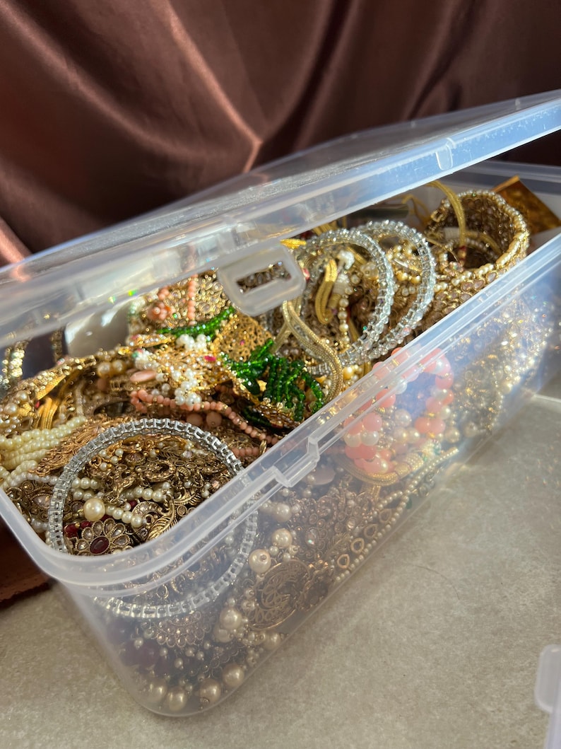 May include: A clear plastic storage container filled with various jewelry pieces. The jewelry includes gold-toned bracelets, necklaces, and earrings, adorned with pearls, beads, and gemstones. The container is partially open, revealing the contents.