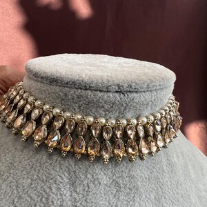 May include: A gold-toned choker necklace with teardrop-shaped, peach-colored gemstones and small white pearls. The necklace is displayed on a gray velvet bust, showcasing its intricate design and elegant style.