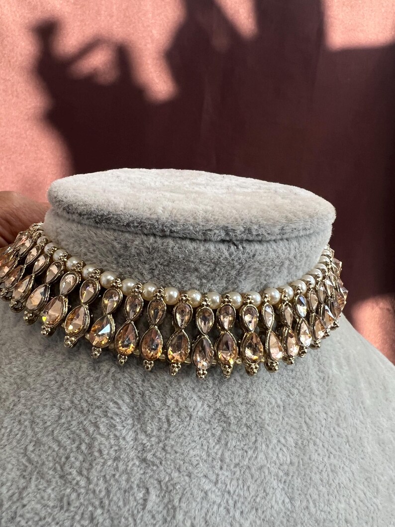 May include: A gold-toned choker necklace with teardrop-shaped, peach-colored gemstones and small white pearls. The necklace is displayed on a gray velvet bust, showcasing its intricate design and elegant style.