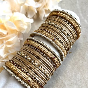 May include: A stack of gold-toned metal bangles featuring intricate textured patterns, embedded with small white faux pearls and tiny red and clear crystals. The bracelets are arranged in a cylindrical formation, showcasing a variety of decorative designs.
