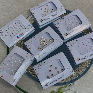 May include: Seven small packs of rhinestones, each with a different colour and design. The packs are white with a gold border and the text "Richie Rich" printed on them.