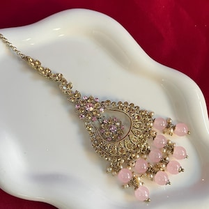 May include: An ornate, gold-toned headpiece with a central teardrop design. The headpiece features pink gemstones, small white pearls, and delicate floral accents. A chain extends from the top for securing the piece. The jewelry is displayed on a white, wavy surface.