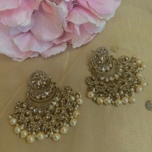 Peut inclure: Une paire de boucles d'oreilles dor&eacute;es avec des accents de perles. Les boucles d'oreilles pr&eacute;sentent un grand motif orn&eacute; avec plusieurs couches de d&eacute;tails complexes. Les perles sont enfil&eacute;es sur un fil et pendent du bas des boucles d'oreilles.