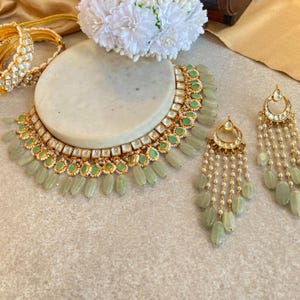 Può includere: Un set di collana e orecchini in oro con pietre verdi e bianche. La collana ha un design a strati con una fila di pietre verdi e una fila di pietre bianche. Gli orecchini sono lunghi e pendenti con pietre verdi e bianche.