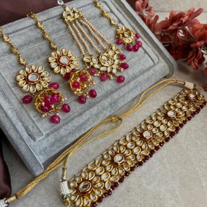 May include: A gold-coloured jewellery set featuring a necklace and headpieces. The necklace and headpieces have a floral design with red and clear accents. The headpieces include dangling red beads and pearls. The jewellery is displayed on a grey surface.