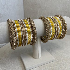 May include: Two sets of gold-toned bangles with yellow and white accents, displayed on a white stand. The bangles feature intricate detailing with small beads and embellishments. The yellow bangles are interspersed with white and pearl-like beads.