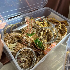 May include: A clear plastic storage container overflowing with various pieces of jewelry. The jewelry includes gold-toned bracelets, necklaces, and earrings, adorned with pearls, beads, and gemstones. A variety of colors are present, including gold, white, pink, and green.