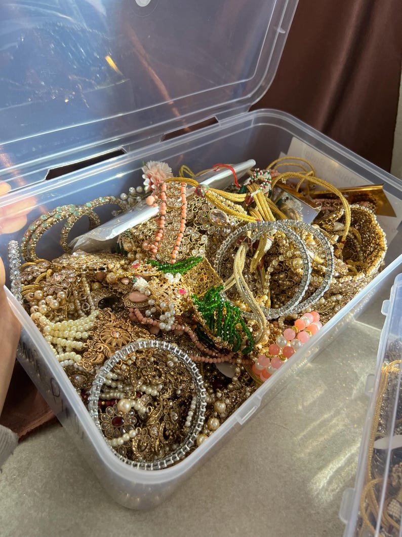 May include: A clear plastic storage container overflowing with various pieces of jewelry. The jewelry includes gold-toned bracelets, necklaces, and earrings, adorned with pearls, beads, and gemstones. A variety of colors are present, including gold, white, pink, and green.