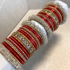 May include: A collection of red and gold bangles with intricate designs and sparkling embellishments. The bangles are stacked together, showcasing a variety of textures and patterns. The jewelry is accented with clear, crystal-like beads.
