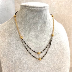 Könnte beinhalten: Eine zweireihige schwarze Perlenkette mit goldenen Akzenten. Die Kette hat kleine, runde goldene Perlen an den Enden und in der Mitte.